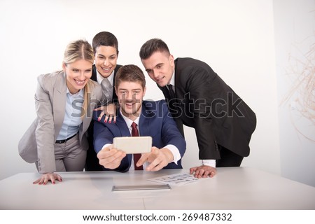 Happy co-workers makes self-ie. Four laughing young business people makes SELF-IE with the sell phone. Office interior, stationery, paper, charts on the table
