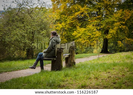 Young woman sitting on a old wooden park bench in the forest during autumn/Young women on park bench/Autumn theme