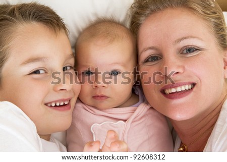 Mother and her kids in the bed, big brother is close to his swaddled sister. All of them smiling happily looking at camera while.
