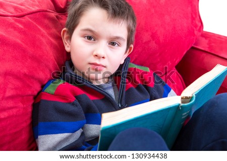 Kid looking at camera while reading a book on the red couch. He has a meaningful look on his face, perhaps distracted.