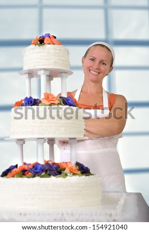 Happy baker lady happily smiling large in front of her cake with her apron and white bandanna, cake has fondant ruffles on the side and decorated with orange and purple gum paste roses