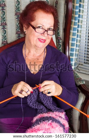 Red haired senior lady looking at you while knitting her purple scarf, she is wearing purple clothing perhaps purple is her favorite color