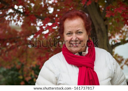 Red hair senior lady looking at you happily and trustfully with her red scarf and white coat under the tree with red falling leaves