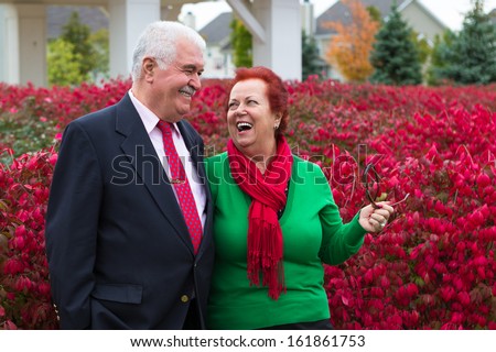 Happy and healthy senior enjoying autumn by the burning bush shrubs, she is wearing Christmas colors, red scarf and green shirt he has suit and tie