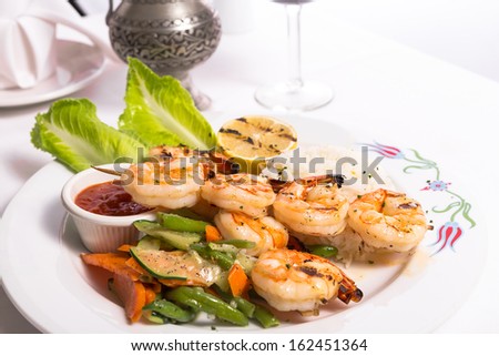 Shrimp skewers are grilled and garnished with veggies and rice served on a white plate along with a silver pitcher