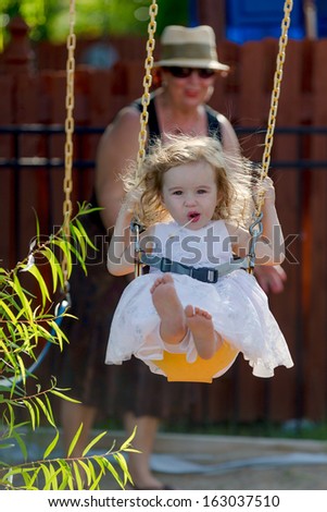 Toddler girl having fun on the swing while having candy with her bare naked feet and pushed by her grandmother with the hat