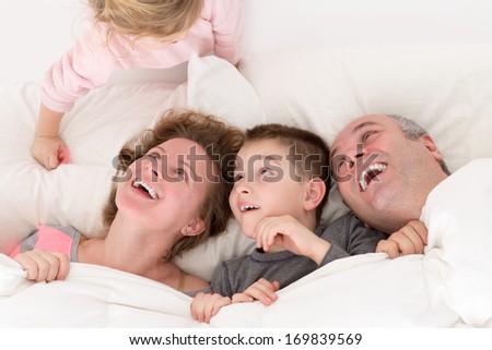 Playful little girl with her family in bed clambering over the top of the pillow making them laugh with amusement and pleasure as her brother and parents snuggle together under the bedclothes