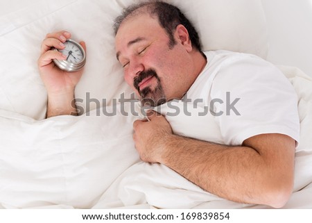 Man Lying In Bed Smiling In Contentment After A Good Nights Sleep Feeling Relaxed And Refreshed As He Struggles To Wake Up In The Morning With His Alarm Clock In His Hand