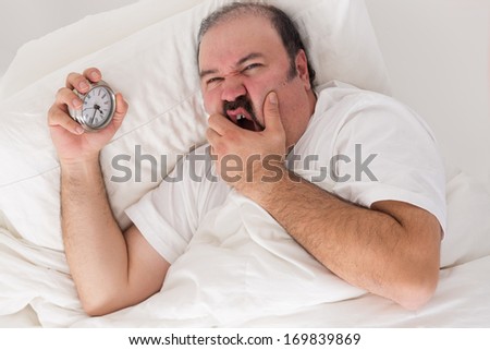 Man suffering from insomnia lying in bed clutching his alarm clock as he checks the time and wonders if he will wake up in the morning for work if he goes to sleep now