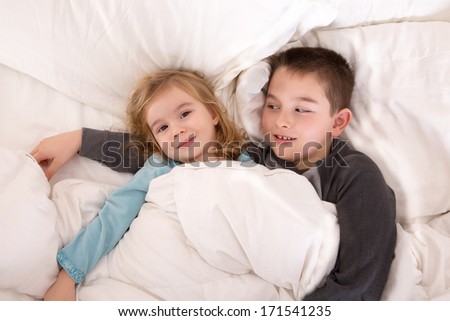 Protective young boy lying in bed with his little sister watching over her as she lies smiling up at the camera as they prepare to go to sleep