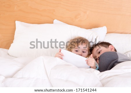 Little brother and sister snuggling up together at bedtime under a warm duvet as they read an e-book or surf the internet on a tablet-pc