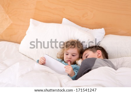 Tired little brother and sister snuggling up together in a warm comfortable bed as they read a bedtime story on a tablet computer before falling asleep