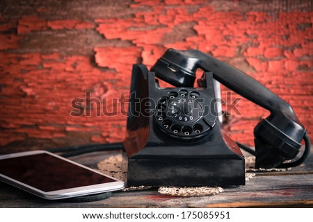 Old rotary telephone with its receiver off the hook alongside a tablet computer showing the old-fashioned and modern forms of communication against a grungy wooden wall with peeling paint