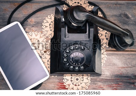 Tablet computer with a blank screen lying alongside a retro rotary telephone with its handset off the hook on a decorative doily, high angle view