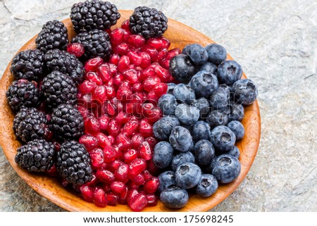 Wooden bowl of fresh blueberries, blackberries and pomegranate seeds arranged in colorful rows, high angle view on an old stone counter top