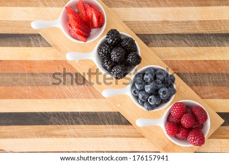 Dishes of fresh berries on decorative striped wood arranged in a diagonal row on a small wooden board including blackberries, raspberries, strawberries and blueberries for a tasty nutritious snack