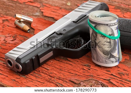 Gun with a roll of 100 dollar banknotes and two bullets lying on a grungy wooden surface with red peeling paint conceptual of crime, robbery, coercion, bribe or mob protection money