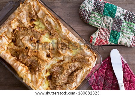 Uncut Turkish borek fresh from the oven with its crisp flaky golden yufka pastry crust standing on a rustic wooden table with oven gloves and a metal spatula, overhead view