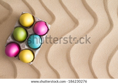 Traditional Easter Egg hunt at the beach with a box of colorful painted eggs hidden on golden sand with a decorative wavy pattern and copy space for your seasonal greeting