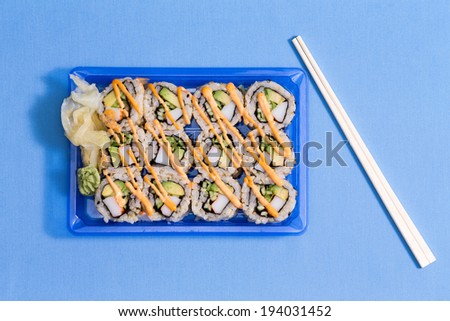 Plastic takeaway punnet of sushi rolls to go with seaweed, raw fish and rice served with traditional Japanese chopsticks, overhead view on blue