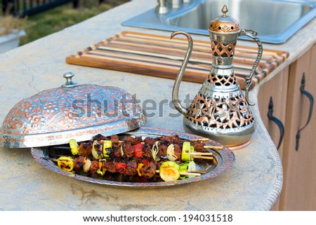 Grilled beef kebabs with tomato, onion and zucchini served on a metal plate with domed cover with a matching elegant pitcher on a concrete counter in an outdoor summer kitchen