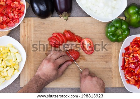 View from above of the hands of a man preparing vegetables in the kitchen slicing tomatoes on a wooden board surrounded by diced zucchini, onion, tomato and whole fresh green bell peppers and eggplant