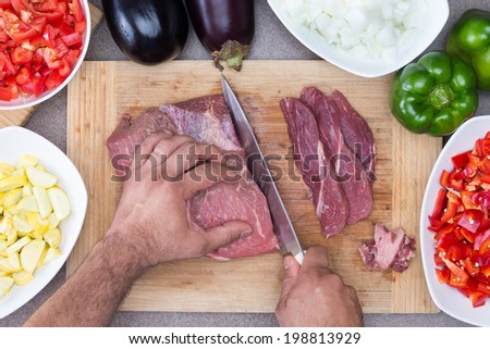 Hands of a man preparing meat and vegetables in a kitchen slicing lean meat on a wooden board with diced zucchini, tomato and onion and whole eggplant and green bell pepper