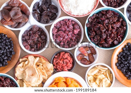 Background pattern and texture of assorted dried fruit displayed in individual round bowls including goji berries, cranberries, apple, coconut, apricot, banana and dates, close up top view on burlap
