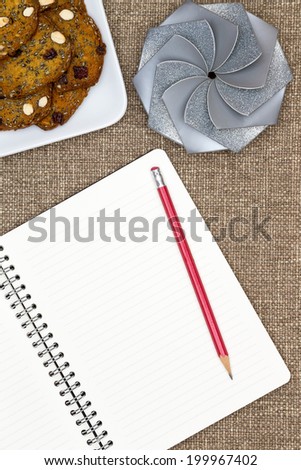 Open blank white notebook with lined pages and a pencil lying on a textured brown cloth with a plate of almond nut cookies and a a decorative round gift with a spiral pattern top