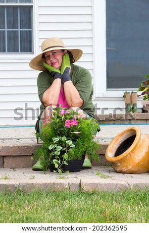 Stylish Grandma in gumboots and sunhat seated on the brick steps of her patio looking pensively at the camera with a lovely smile as she sits potting up houseplants in flowerpots to decorate the house