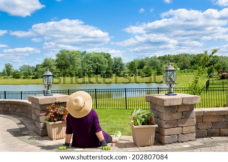 Senior lady gardener taking a break to admire the view from her front patio over a lake and lush green countryside sitting with her back to the camera in a straw sunhat