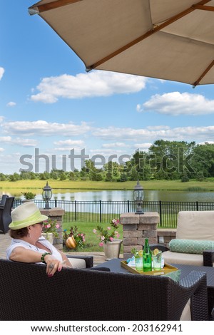 Rear view of a woman relaxing on an outdoor patio enjoying the view over a calm peaceful lake and lush countryside as she enjoys a glass of iced bottled water on a hot summer day