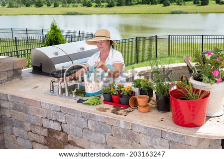 Senior lady filling a watering can at a sink in a summer kitchen on an outdoor patio to water her collection of potted plants and houseplants standing ready on the counter with a lake backdrop