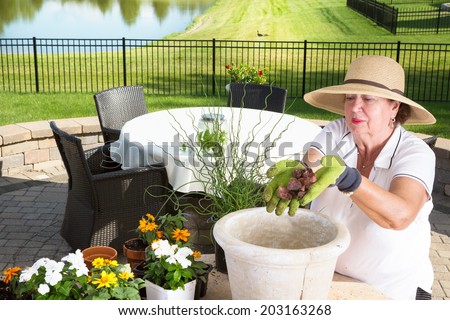 Senior lady gardener potting up a large planter on an outdoor brick patio adding a handful of stones to the bottom for drainage and aeration before adding the plants and soil