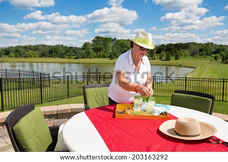 Senior lady preparing iced drinks outdoors adding fresh mint and lemon to glasses on a tray on a garden table with red cloth overlooking a tranquil lake and lush countryside