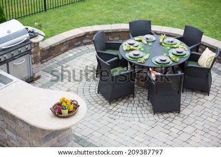 Elegant outdoor living space on a paved brick patio with a summer kitchen and barbecue and a table laid with formal place settings for dinner, high angle view