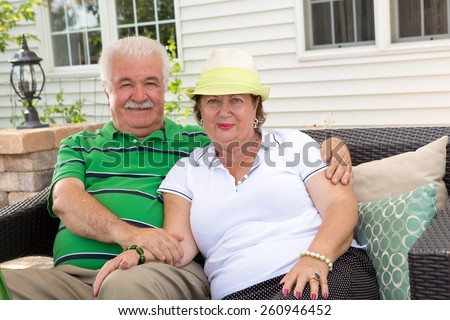 Affectionate elderly couple relaxing on an outdoor patio sitting arm in arm on a garden sofa in front of their house smiling at the camera