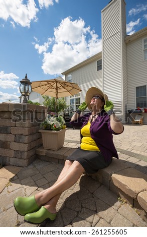 Elderly woman in gardening clothes and a sunhat sitting on the steps of her outdoor patio with her head tilted back enjoying the spring sunshine - spring is here