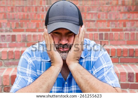 Boredom - the enemy of human happiness in a conceptual image of a middle-aged man with a goatee sitting resting his chin in his hands with a glum expression staring at the camera against a brick wall