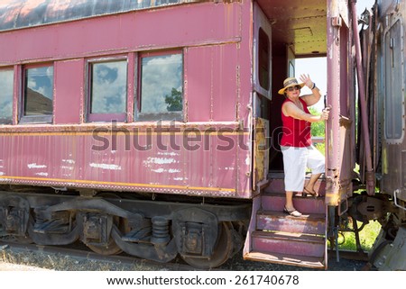 Elderly woman in trendy summer clothing standing waving at the camera from the steps of a vintage train carriage in a conceptual image of travel and vacations