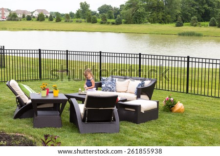Cute little blond girl standing amongst comfortable modern design garden furniture with armchairs and tables arranged on a neat green lawn overlooking a tranquil pond and wrought iron railing