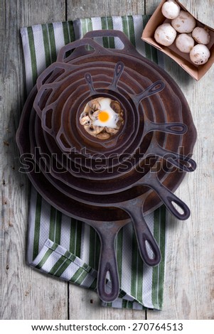 Sunny side up fried egg and mushrooms in a nested stack of old cast iron frying pans in a rustic kitchen, overhead view