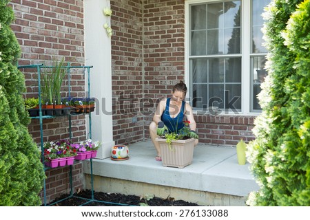 Woman tending to newly potted ornamental spring plants on her patio kneeling alongside a large flowerpot on a cement patio in front of the brick wall of her home, view between two cypress trees
