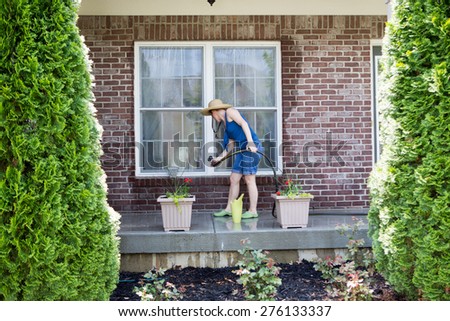 Woman washing the exterior windows of a house with an attachment on a hose as she cleans and refreshes the house after winter for the new spring season, view framed by two evergreen cypresses