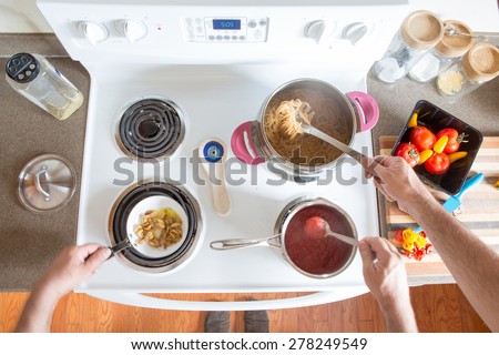 Two men preparing healthy brown wholewheat spaghetti cooking together at the hob as a team stirring the boiling pasta and sauce, overhead view of their hands and ingredients