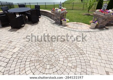 Ornamental brick paved outdoor patio with a circular design in the bricks with dining furniture and colorful flowers in flowerpots flanking steps and an entrance, view from above after rain