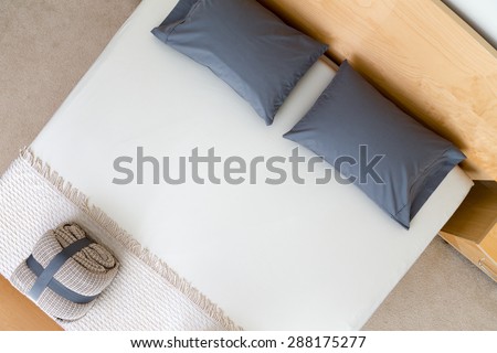 Overhead view of a neat king size bed in a hotel or house with black pillows on a white counterpane with a folded and strapped rug at the food and a wooden headboard