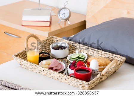Close Up of Breakfast Tray Filled with Variety of Foods Sitting on Unmade Bed Next to Bedside Table with Lamp and Alarm Clock in Hotel Room
