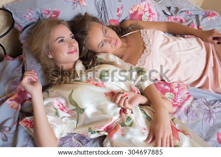 blond sisters or sexy girl friends having fun relaxing in pajamas lying on white bed looking at camera