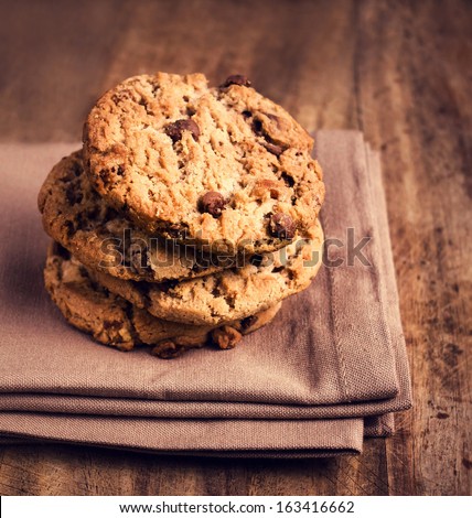 Stacked chocolate chip cookies on brown cloth over wooden background in country style, macro, brown tones color